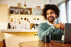 Smiling male sitting at his dining table with a drink and his laptop. Looking away. Kitchen in the background.
