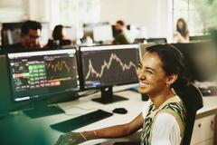 Woman smiling while sitting behind her desk, screens displaying financial information.