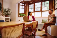 Female sitting at a table with laptop talking to male with coffee mug at home

