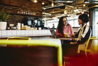 Two woman sitting in restaurant having a converstation, looking at laptop, smiling.