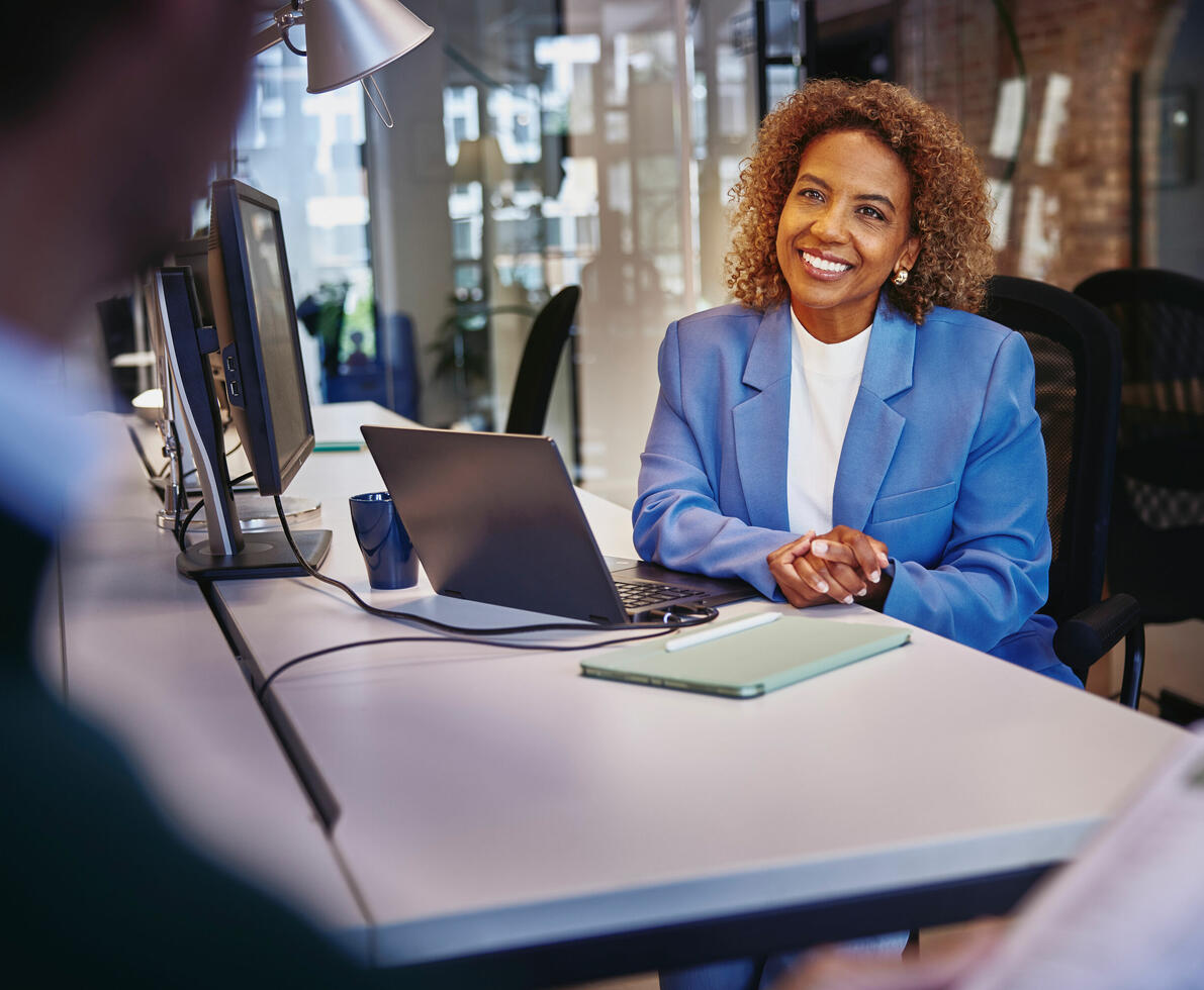 Female with blue blazer sitting behind computer at a desk, smiling to a person Female with blue blazer sitting behind computer at a desk, smiling to a person