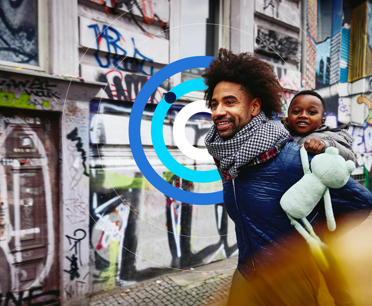Homem sorridente com uma criança às costas a passear numa cidade.