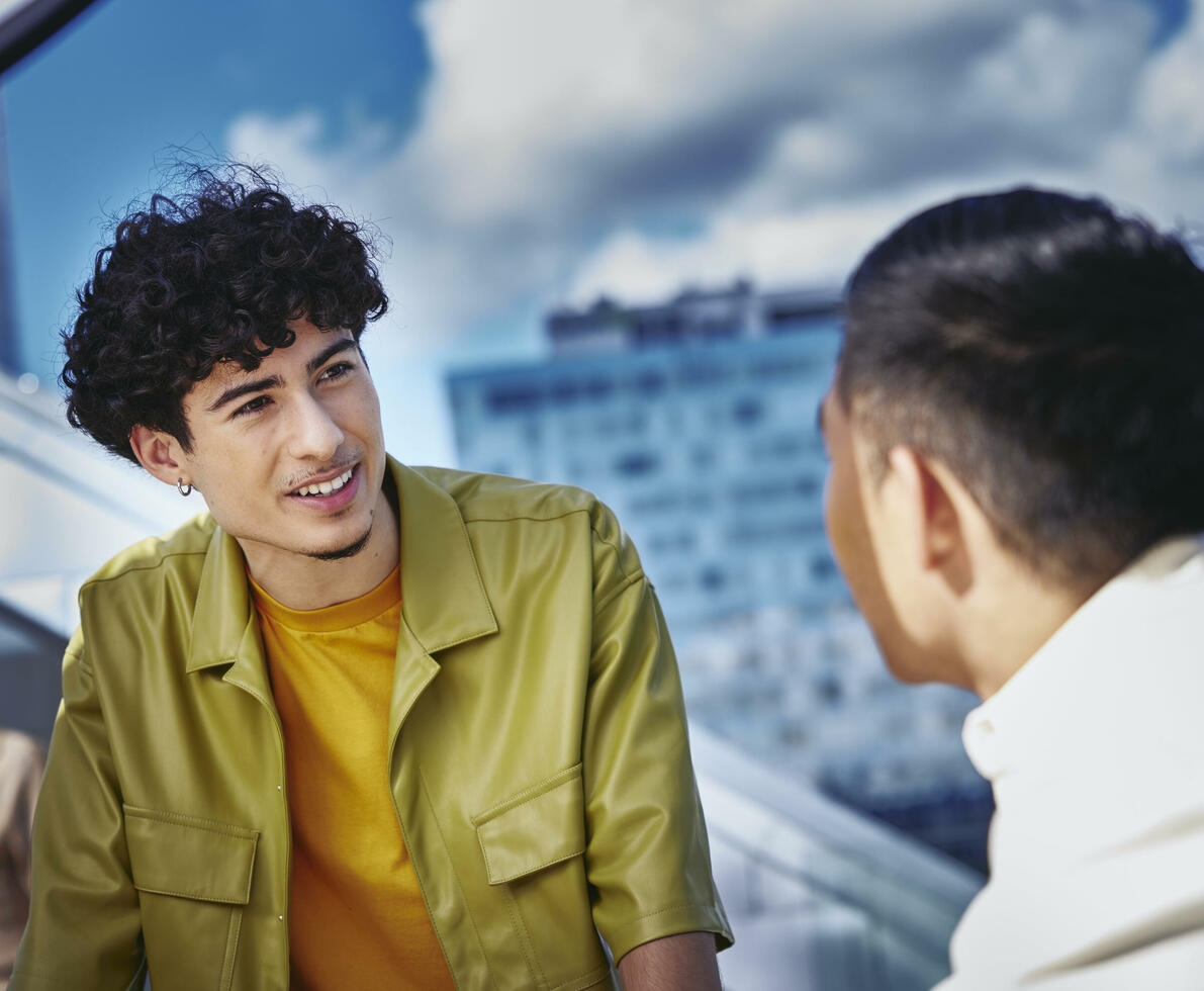 Male in yellow shirt and male, standing outside on a balcony, talking