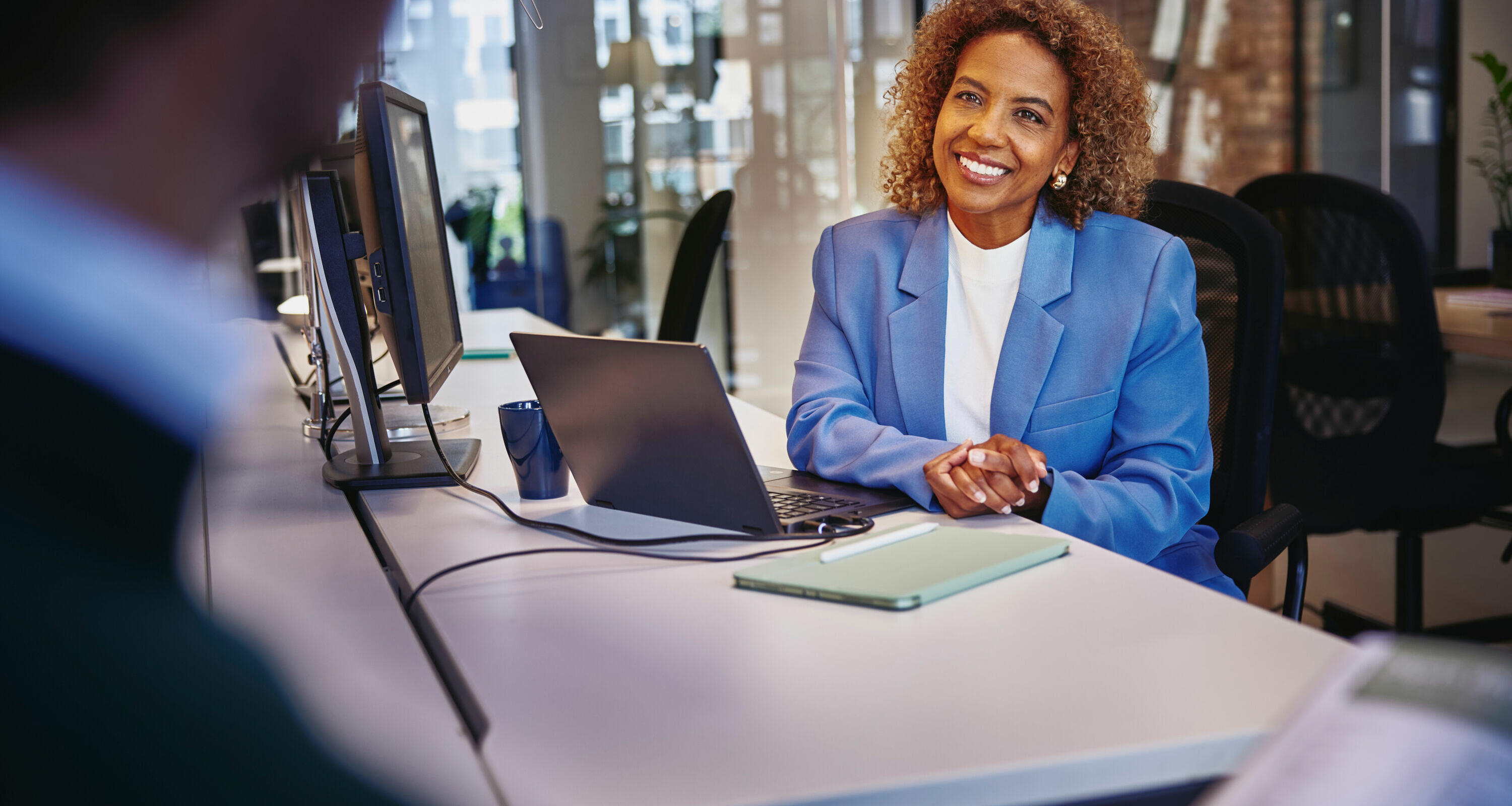 Female with blue blazer sitting behind computer at a desk, smiling to a person
